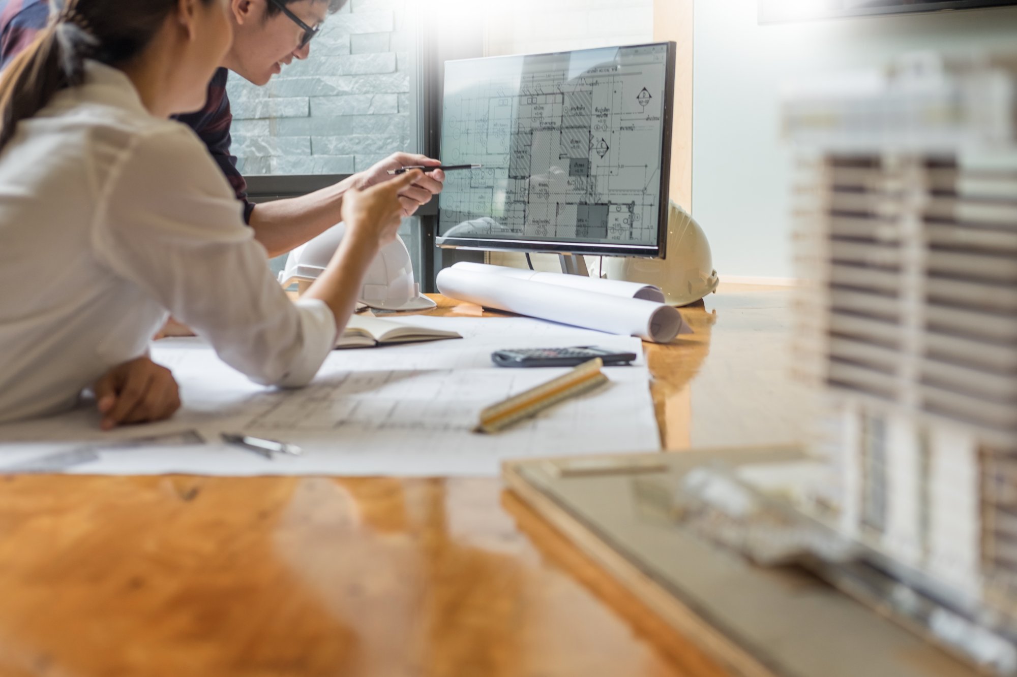 Engineers reviewing technical drawings on screen with blueprints spread across drafting desk